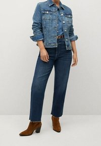 Chaqueta de mezclilla en azul medio con cierre de botones, dos bolsillos frontales y mangas enrolladas, combinada con unos jeans oscuros de cintura alta y botas de tacón marrones.