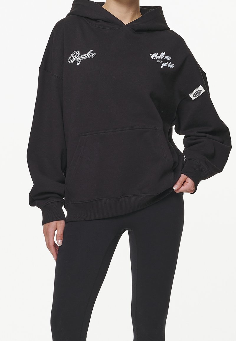 Personne portant un sweat à capuche noir ample avec texte blanc "Regular" et "Call me if you get lost", associé à des leggings noirs sur fond blanc.