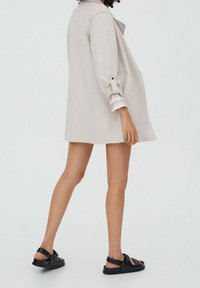 Manteau beige oversize avec un seul bouton, manches retroussées et coupe ample. Sandales noires avec des brides texturées et un dos ouvert.