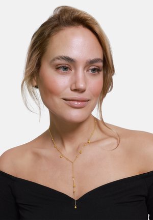 Guld lariat halskæde med runde og ovale perler, der har et delikat kæde-design. Bæres på bare skuldre med en off-shoulder top.