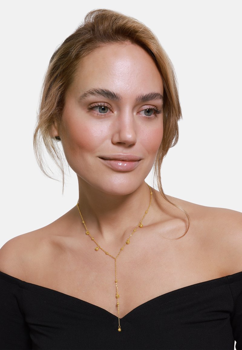 Collana lariat dorata con perle rotonde e ovali, caratterizzata da un design di catena delicato. Indossata su spalle scoperte con un top a spalle scoperte.