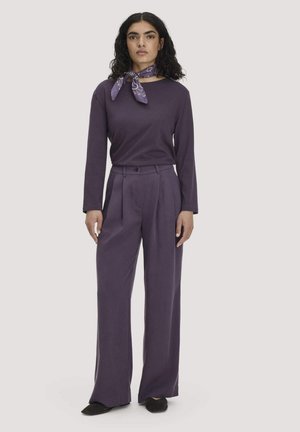 Vrouw met krullend haar die een paarse top met lange mouwen draagt, wijde paarse broek, zwarte schoenen en een paarse, met patroon, halsdoek.