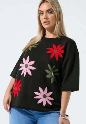 Mujer que lleva un suéter negro con grandes patrones florales en rojo, rosa y verde combinado con vaqueros azul claro y una pulsera de plata.