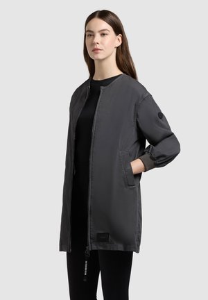 Frau trägt eine lange, dunkelgraue Jacke mit Reißverschluss und Taschen, steht mit einer Hand in der Tasche vor einem einfarbigen hellgrauen Hintergrund.