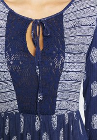 Robe bleu marine avec motifs blanc paisley et géométriques, présentant un col en panneau de dentelle noué par un ruban à l'avant.