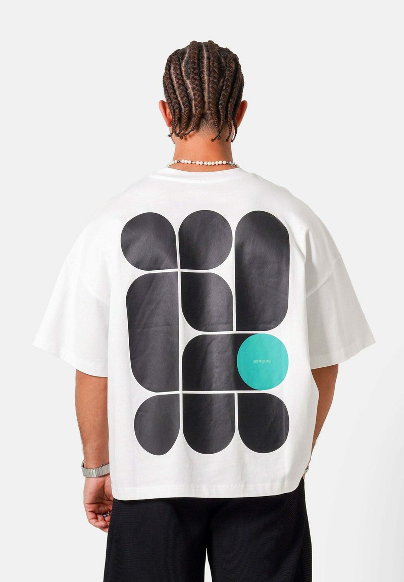 Persona con cabello trenzado que lleva una camisa blanca con grandes formas abstractas negras y un círculo azul verdoso en la espalda, de pie frente a un fondo liso.