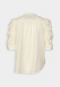 Blouse crème à manches courtes bouffantes, motif à pois métalliques dorés, et détail de soutache au dos.