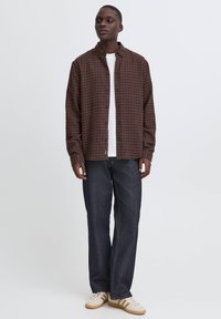 Homme portant une chemise à carreaux marron boutonnée par-dessus un t-shirt blanc, un jean bleu foncé, et des baskets blanches avec des bandes beige, debout sur un fond blanc.