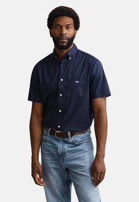 Man with beard wearing dark blue short-sleeve button-up shirt and light blue jeans, standing against a plain background.