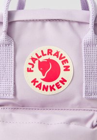 Lila szövet hátizsák, kerek, krémszínű logóval, amelyen egy vörös róka dizájn látható, valamint a "FJÄLLRÄVEN KÄNKEN" felirat. Kiegészítők között szőtt pántok szerepelnek.