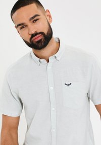 Camisa de botones de manga corta en gris claro, hecha de tela tejida, con un bolsillo que presenta un diseño oscuro bordado. Textura suave y ajuste casual.