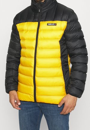 Homme portant une veste matelassée jaune et noire avec fermeture éclair à l'avant et écusson logo sur la poitrine, assortie à un jean bleu.
