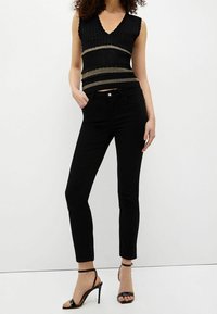 Mujer vestida con un top de punto sin mangas negro ajustado con rayas metálicas, jeans negros ajustados y sandalias de tacón alto negras.