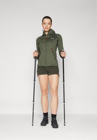 Chaqueta deportiva verde con acentos naranjas, bolsillos con cremallera y capucha ajustable; combinada con pantalones cortos verdes y calzado negro, sosteniendo bastones de senderismo.