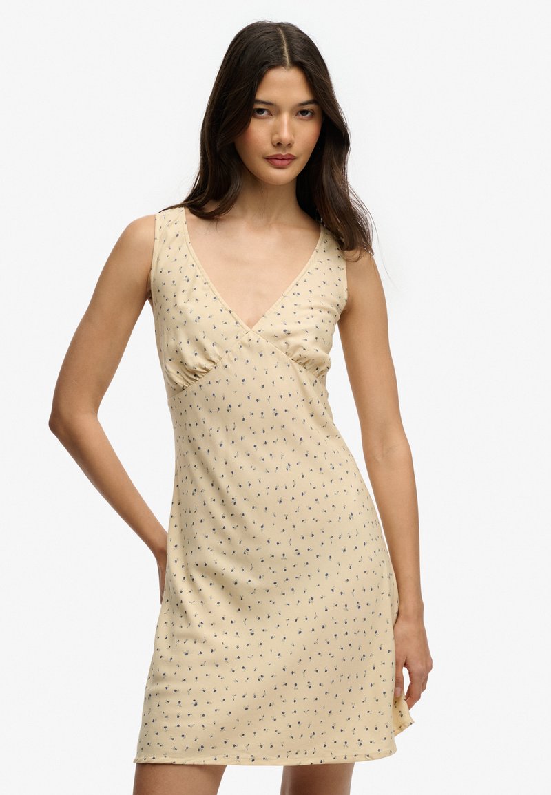 Jeune femme aux longs cheveux foncés portant une robe sans manches jaune pâle avec un petit imprimé floral bleu, debout devant un fond blanc uni.