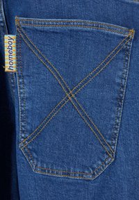 Denim-kangas, jolla on sininen sävy. Takataskussa on oranssit ompeleet 'X'-kuviossa. Kiinnitettynä on lappu, jossa on "homeboy" raidallisissa väreissä.