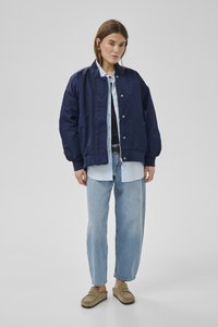 Junge Frau trägt eine marineblaue Bomberjacke, ein hellblaues Hemd mit Knöpfen, hellblaue Jeans und beige Slipper, steht vor einem schlichten grauen Hintergrund.