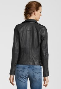 Schwarze Motorradlederjacke mit Schulterklappen, vertikalen Nähten und tailliertem Schnitt, kombiniert mit blauen Jeans mit aufgenähten Hintertaschen.