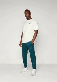 Kremo spalvos trumpomis rankovėmis marškinėliai su krūtinės kišene, derinami su žaliais džemperiais su juostelėmis ir Adidas logotipu, dėvimi su baltais sportbačiais.