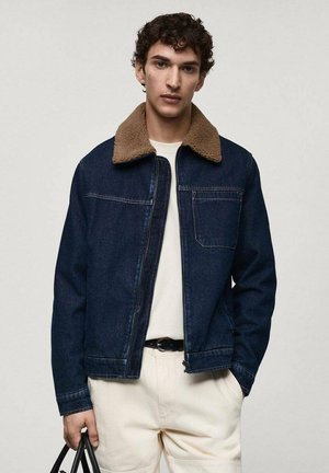 Veste en denim avec un col en faux fourrure marron. Couleur bleu foncé avec des coutures visibles et une poche avant. Portée sur une chemise blanche et un pantalon clair.