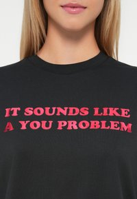Prim-plan al unei femei care poartă un tricou negru cu un text îndrăzneț în roșu care spune „AUDIO CA O PROBLEMĂ A TA.”