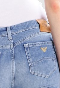 Jasnoniebieskie jeansy z denimu z brązową skórzaną metką na pasie i złotym emblematem na tylnej kieszeni. Charakteryzują się klasycznym krojem i przeszyciami.