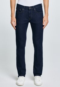 Bărbat îmbrăcat în blugi slim fit bleumarin și adidași albi, stând pe un fundal simplu.