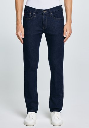 Hombre con jeans ajustados azul oscuro y zapatillas blancas de pie frente a un fondo liso.