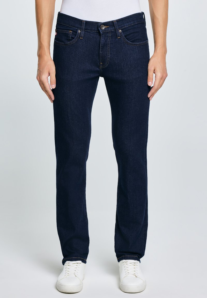 Bărbat îmbrăcat în blugi slim fit bleumarin și adidași albi, stând pe un fundal simplu.