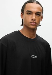 Joven con el pelo trenzado que lleva una camiseta negra con el texto "Le Petit Café" en el frente, contra un fondo liso claro.