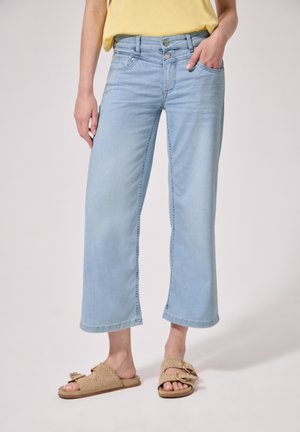 Lyseblå jeans med vidde ben og dobbelt knaplukning, båret med en gul top og beige åbne sandaler med nitter.