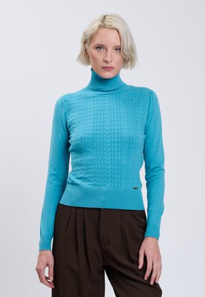 Vrouw met kort blond haar draagt een felblauwe kabelgebreide coltrui en een bruine broek, staand tegen een egale lichte achtergrond.