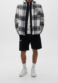 Koszula w kratę w szaro-białe wzory, zestawiona z czarnym t-shirtem i czarnymi szortami cargo. Białe sneakersy i szare skarpetki dopełniają stylizację.