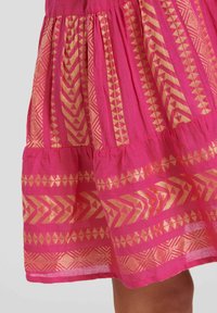 Rosa bomullskjol med gyllene geometriska mönster. Har samlade lager och en utsvängd form. Tyget har en mjuk, texturerad yta.