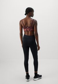 Frau mit geflochtenem Haar, die einen kastanienbraunen Sport-BH, schwarze hochgeschnittene Leggings, schwarze Socken und schwarze Laufschuhe trägt und vor einer weißen Wand steht.