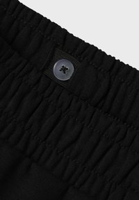 Cintura elástica negra con textura fruncida, con un botón negro redondo en la parte superior. Tejido suave con un ligero estiramiento.