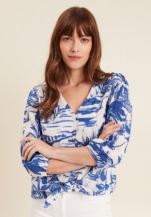 Blusa blu e bianca a fantasia con scollo a V, dettagli con bottoni e maniche a tre quarti. Il tessuto ha un aspetto testurizzato e un fronte annodato.