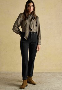 Čierne nohavice s vysokým pásom a priamym strihom, doplnené o košeľu v hnedej kárované vzore s viazačkou na krku. Hnedé semišové členkové čižmy.