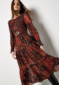Długa, zwiewna sukienka z ciemnym, kwiatowym wzorem w odcieniach czerwieni, brązu i czerni. Ma długie rękawy, paski w talii i warstwowy dół.