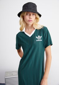 Zaļš sporta kleita ar baltu apkakli, kurā ir vertikālas svītras un Adidas logo. Melns kabatas cepure ar ziedu rakstu un logo.