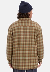 Braun-gelb-blau kariertes Flanellhemd mit geknöpftem Kragen, langen Ärmeln und geradem Saum. Getragen mit einer dunklen Mütze.