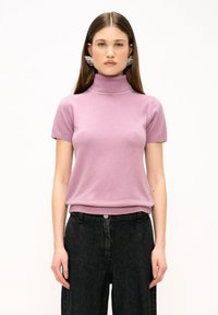 Maglione rosa chiaro a maniche corte con colletto a dolce vita, realizzato in un tessuto morbido e con bordi a costine. Abbinato a jeans scuri a vita alta.