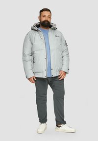 Lichtgrijze pufferjack met capuchon, zwarte accenten en een rits aan de voorkant. Draag het over een blauwe shirt, gecombineerd met donkere broeken en witte sneakers.