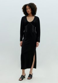 Schwarzes langärmliges Kleid mit tiefem Ausschnitt, dekorativer silberner Naht und seitlichem Schlitz. Kombiniert mit schwarzen Wedge-Sandalen.