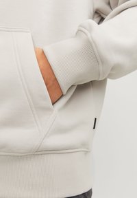 Sweat-shirt gris clair avec une poche kangourou, des poignets et un ourlet côtelés. Le matériau semble doux avec une texture lisse. Étiquette de marque discrète visible.