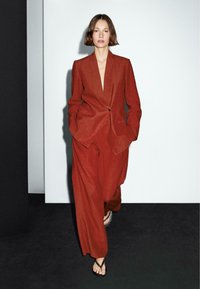 Rust-colored suit with a fitted blazer and wide-leg trousers, made of textured fabric. Open front with a single button, and flip-flops.