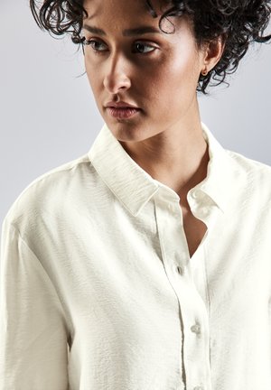 Camisa de color crema, abotonada, con una tela texturizada, que presenta un cuello tipo banda y sutiles arrugas ligeras para un detalle adicional.