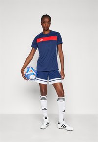 Marineblauw voetbaluniform met rode streep en witte afwerking; inclusief shorts, kniekousen en een voetbal met kleurrijk ontwerp, vastgehouden door een speler.