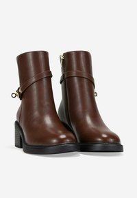 Botas de tobillo de cuero marrón con puntera redondeada, tacón apilado, cremallera lateral y una correa decorativa con un acento dorado. Textura suave.