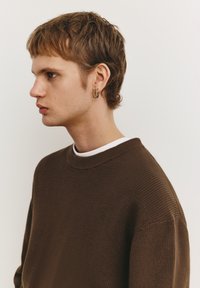 Barna kötött pulóver bordázott textúrával, kerek nyakkivágással és réteges fehér pólóval. A személy rövid hajú és karika fülbevalót visel.